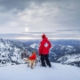 A man and a dog stand on the top of a snowy mountain. The dog is wearing the Ruffwear Vert Jacket